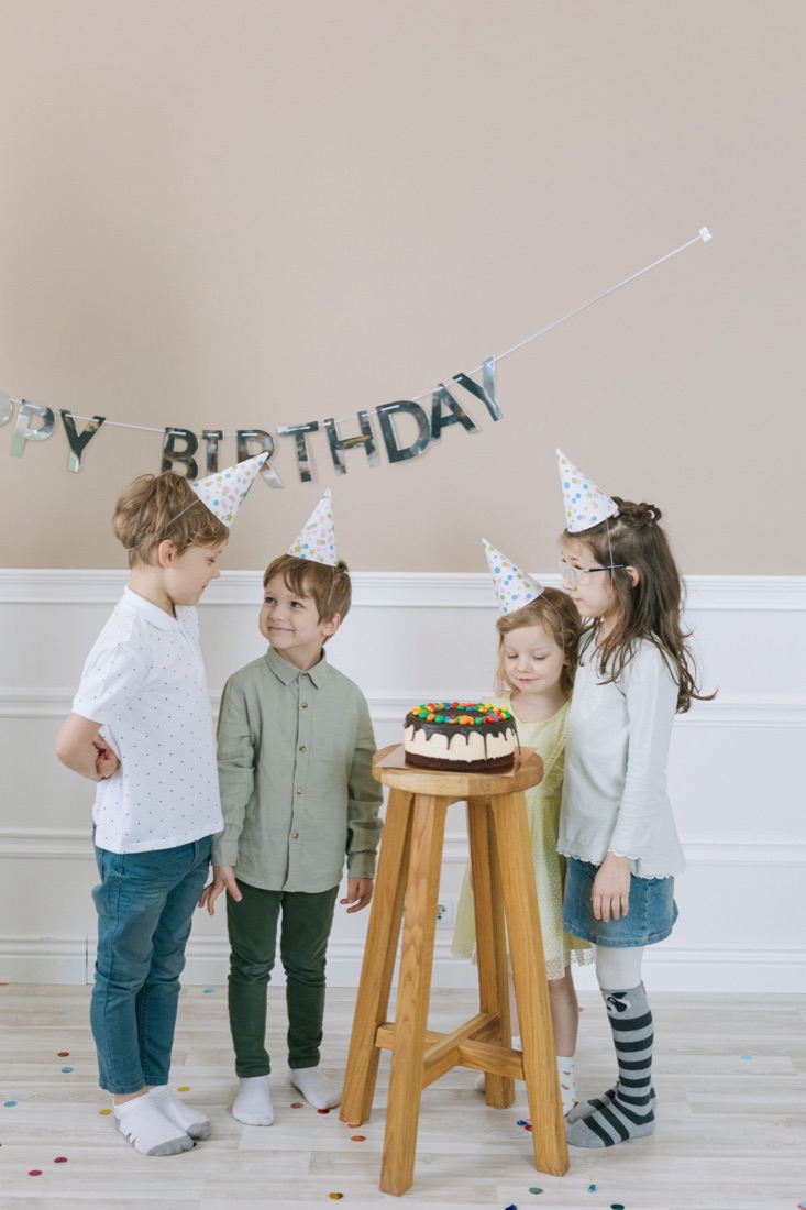 Four children in polka-dot party hats round a chocolate cake under a Happy Birthday garland.