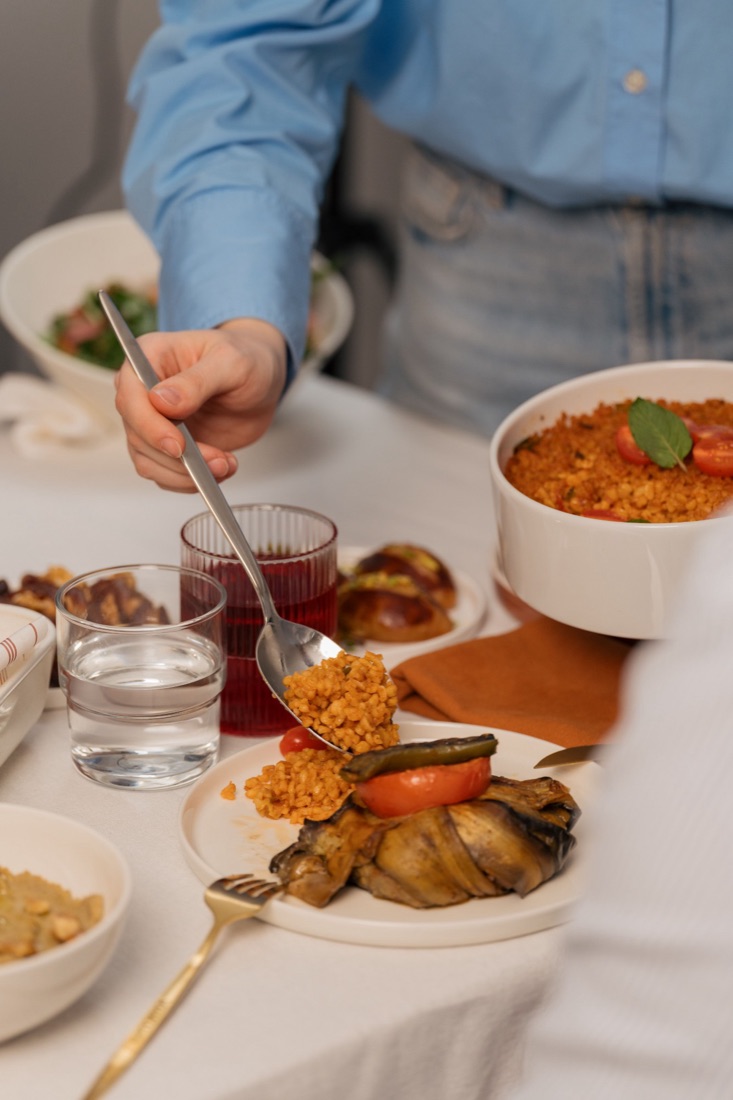 Someone in a blue shirt spoons bulgur pilaf onto a plate beside a stuffed aubergine.