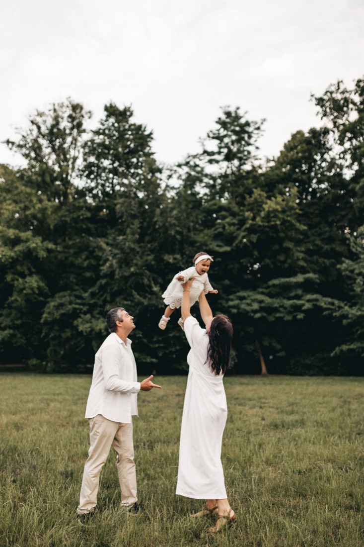 A family dressed in white in a field; a woman lifts a laughing baby into the air.
