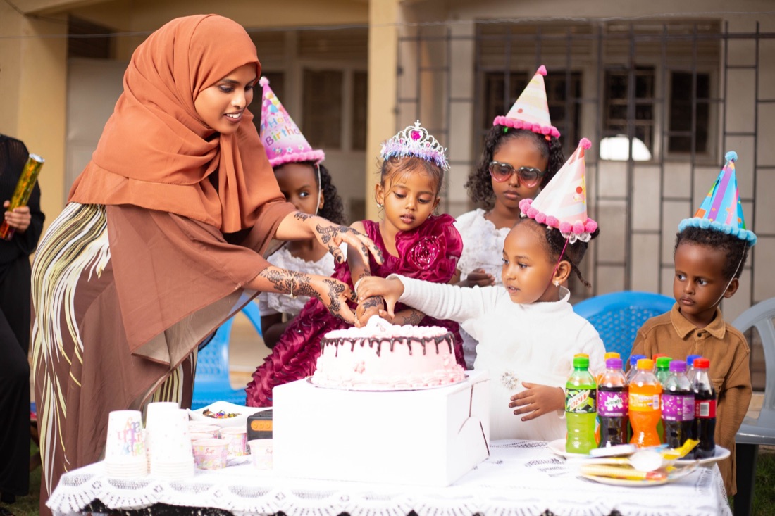 A woman in a rust hijab helps children in party hats and a tiara cut a pink birthday cake outdoors.